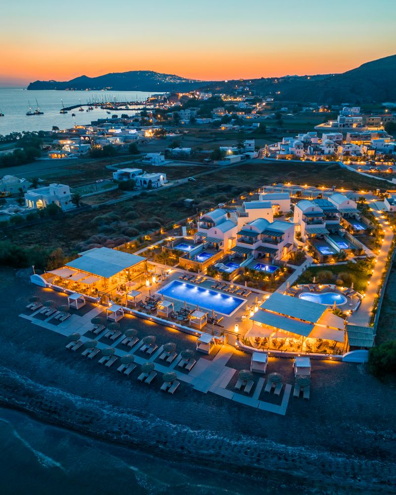 Aerial night view of Sea Breeze Santorini Beach Resort illuminated at sunset, with pools, beachfront terraces and the marina of Vlychada in the background
